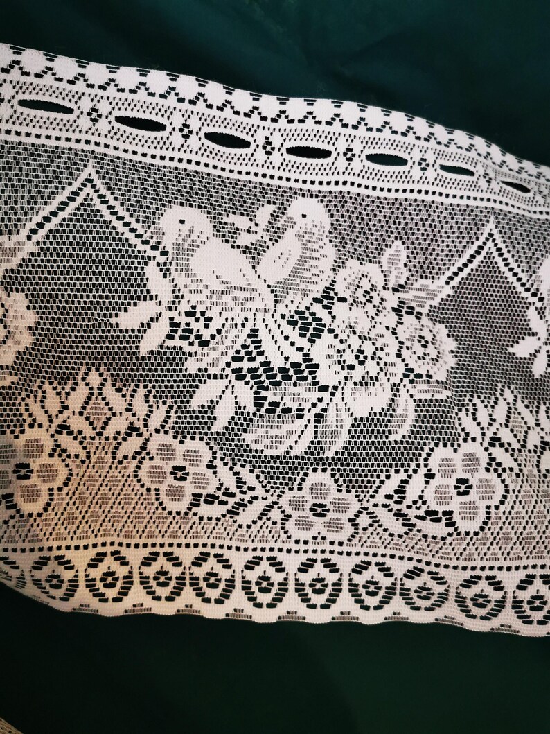 May include: White lace fabric with a floral and bird pattern. The lace features a detailed design with flowers, leaves, and birds, creating an intricate and delicate texture. The fabric is likely used for decorative purposes.