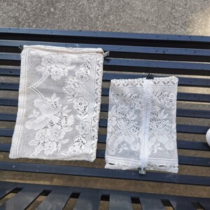 May include: Two white lace pouches with floral and bird patterns. The pouches have zippers and are rectangular. The larger pouch is on the left, and the smaller one is on the right. The pouches are on a dark blue metal bench.