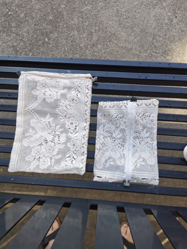 May include: Two white lace pouches with floral and bird patterns. The pouches have zippers and are rectangular. The larger pouch is on the left, and the smaller one is on the right. The pouches are on a dark blue metal bench.