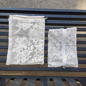 May include: Two white lace pouches with floral patterns and zippers. The larger pouch has a tan zipper, while the smaller one has a white zipper. Both pouches are rectangular and rest on a dark blue metal bench.