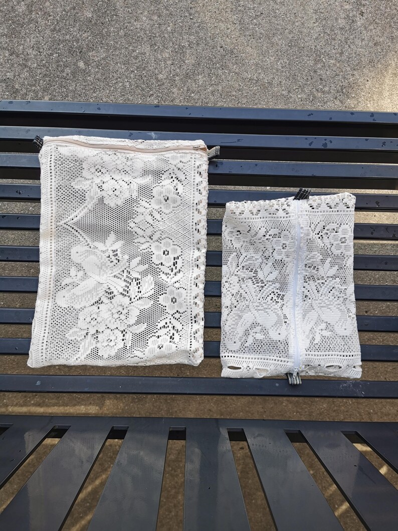 May include: Two white lace pouches with floral patterns and zippers. The larger pouch has a tan zipper, while the smaller one has a white zipper. Both pouches are rectangular and rest on a dark blue metal bench.