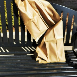 Puede incluir: Una bolsa de papel marrón doblada descansa sobre un banco de metal negro. La bolsa está arrugada y parece vacía. El banco tiene un diseño de listones y está situado al aire libre, con la luz del sol proyectando sombras.