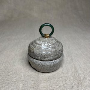 Puede incluir: Una caja de joyería de piedra gris con un asa de anillo verde oscuro asegurada con alambre dorado. La caja es redonda con tapa y base, y se asienta sobre una superficie de color neutro. La piedra tiene un aspecto texturizado.