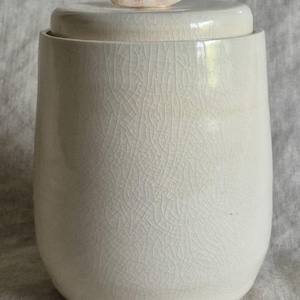 May include: A cylindrical ceramic jar with a cream-colored crackle glaze finish. The jar features a matching lid topped with a single, smooth, white seashell handle. The surface of the container displays a fine network of thin, dark lines.