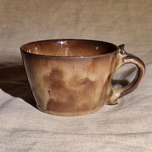 May include: A handcrafted ceramic coffee cup with a distinctive handle. The cup showcases a mix of brown and tan glazes, giving it a rustic, earthy appearance. The inside is a darker brown, and the cup has a smooth, glossy finish.