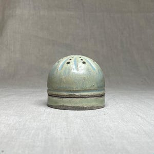 以下が含まれることがあります： ドーム型のトップと円筒形のベースを備えた、手作りのセラミックソルトシェーカー。シェーカーは緑と青の色合いで釉薬がかけられており、塩を出すための小さな穴が上部にあります。デザインは素朴で有機的です。