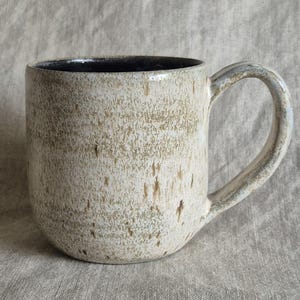 May include: A ceramic mug with a rounded body and a curved handle. The exterior features a speckled, textured finish in shades of cream, beige, and light brown. The interior of the mug is a solid, dark charcoal black color with a smooth finish.