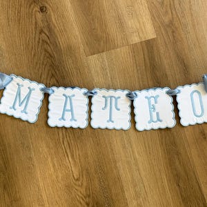 May include: A light blue banner spelling "MATEO" with white scalloped-edge lettered panels. The banner is strung with light blue ribbon and has bows at each end. The banner is displayed on a wooden surface.