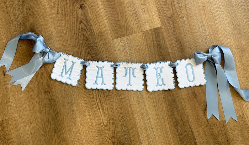 May include: A light blue banner spelling "MATEO" with white scalloped-edge lettered panels. The banner is strung with light blue ribbon and has bows at each end. The banner is displayed on a wooden surface.