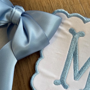 May include: A light blue satin ribbon bow next to a white, scalloped-edge fabric with a light blue embroidered letter "M". The bow is tied and has a smooth, shiny texture. The fabric has a soft, plush appearance.