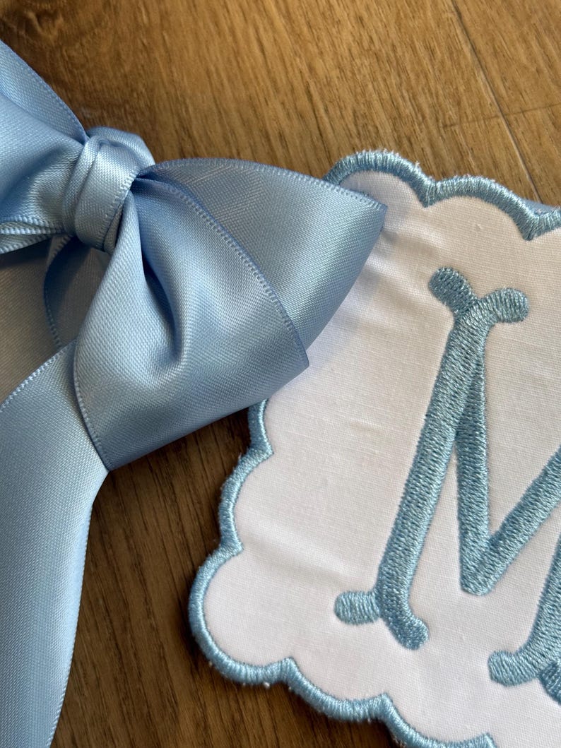 May include: A light blue satin ribbon bow next to a white, scalloped-edge fabric with a light blue embroidered letter "M". The bow is tied and has a smooth, shiny texture. The fabric has a soft, plush appearance.
