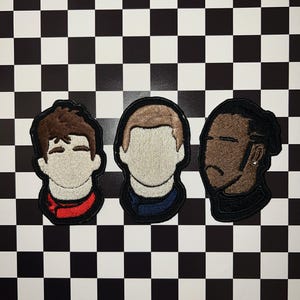 Peut inclure: Trois patchs brodés représentant des portraits de personnes en dessin animé. Les patchs sont noirs avec des accents bruns, rouges et bleus. Les patchs sont sur un fond à carreaux noir et blanc.