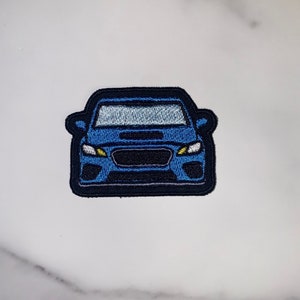 Op de afbeelding: Een blauwe geborduurde patch met een auto, de voorkant van het voertuig is zichtbaar. De koplampen zijn geel, de grille is zwart en de auto heeft een zwarte omtrek.