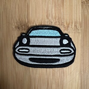 May include: Embroidered patch depicting the front of a car. The patch has a light grey body, black trim, and light blue accents for the windscreen. The headlights are white circles. The patch is on a light brown wooden surface.