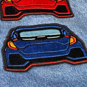 Könnte beinhalten: Zwei gestickte Aufnäher von Autos, eines rot und eines blau, auf einem Jeansstoff-Hintergrund. Die Autos sind in die gleiche Richtung ausgerichtet und haben schwarze Umrisse.