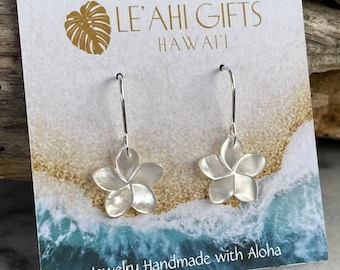 May include: A pair of silver earrings with white shell plumeria flower charms. The earrings are displayed on a white card with the text "LE'AHI GIFTS HAWAII" and "Jewelry Handmade with Aloha".