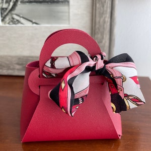 May include: A small, burgundy-colored faux leather handbag with a top handle and a black and pink patterned scarf tied around the handle.
