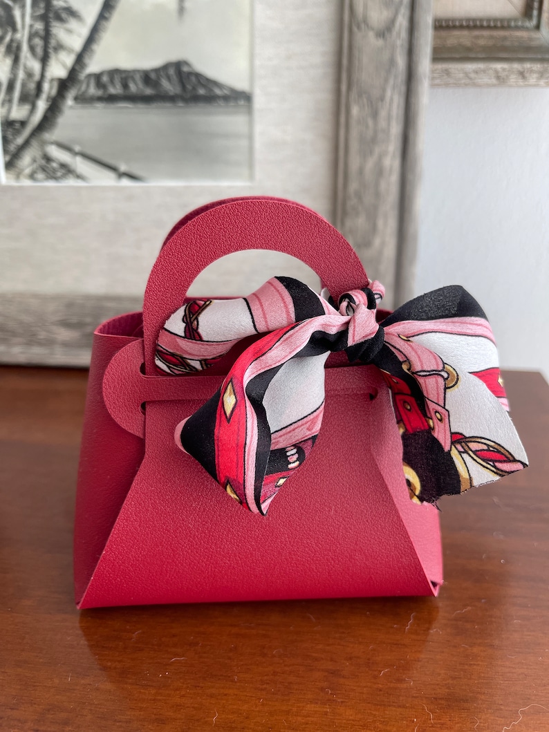 May include: A small, burgundy-colored faux leather handbag with a top handle and a black and pink patterned scarf tied around the handle.
