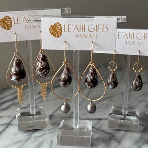 May include: Earrings on display. Gold-toned wire earrings with teardrop-shaped shells and dangling chains or gray pearls. The shells are brown with white patterns. The display cards read "LE'AHI GIFTS HAWAII".