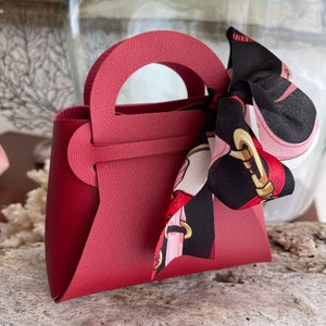 May include: A small, burgundy-colored faux leather handbag with a top handle and a black and pink patterned ribbon tied around the handle.