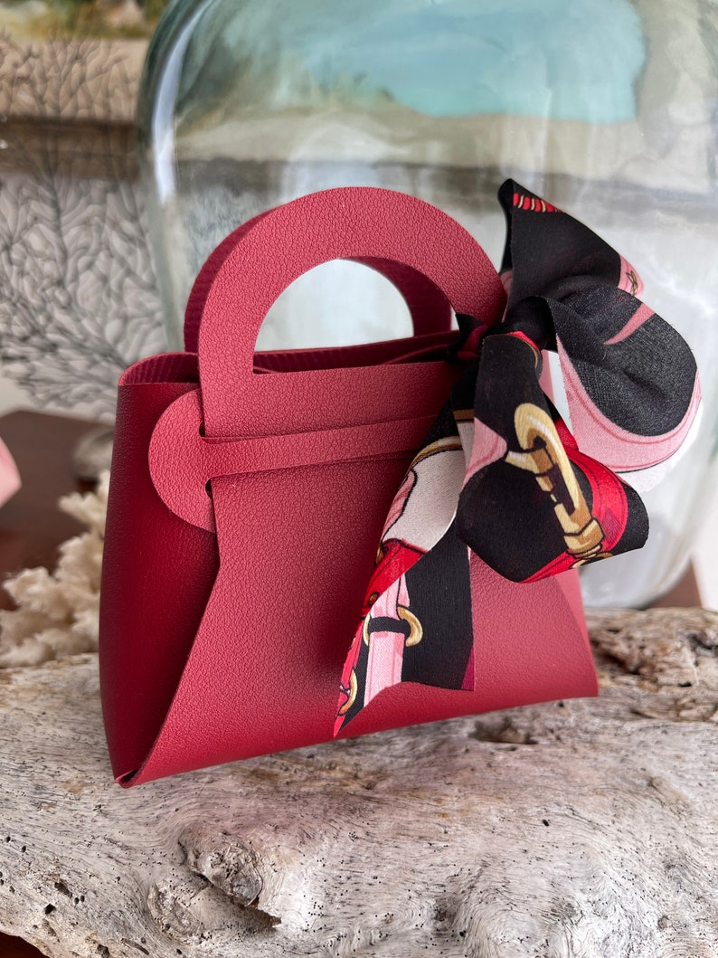 May include: A small, burgundy-colored faux leather handbag with a top handle and a black and pink patterned ribbon tied around the handle.