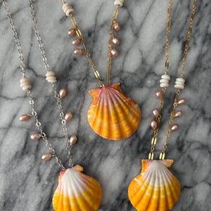 May include: Three necklaces featuring vibrant orange and yellow scallop shell pendants. Two necklaces have gold-tone chains with small pink pearls and white disc beads, while one necklace features a silver-tone chain with pink pearls and white beads.