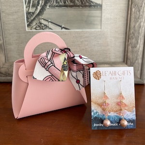 May include: A pink faux leather handbag with a pink and white patterned scarf. The bag is next to a card with the text "Le'ahi Gifts Hawaii" and a pair of pink and gold earrings.