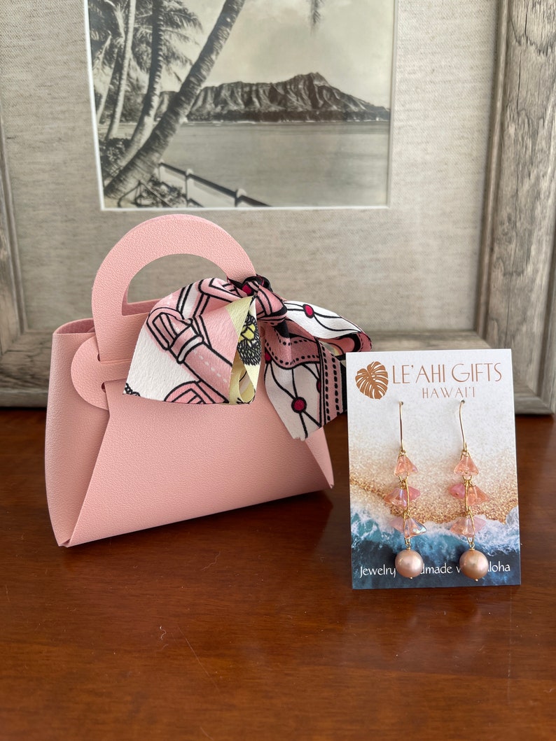 May include: A pink faux leather handbag with a pink and white patterned scarf. The bag is next to a card with the text "Le'ahi Gifts Hawaii" and a pair of pink and gold earrings.