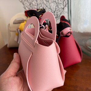 May include: Three small, colorful, faux leather handbags with top handles. The pink handbag is in focus and has a unique heart-shaped handle.