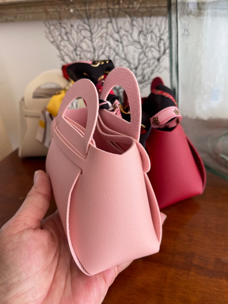 May include: Three small, colorful, faux leather handbags with top handles. The pink handbag is in focus and has a unique heart-shaped handle.