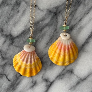 Peut inclure: Deux colliers coquillages avec des chaînes dorées. Chaque collier présente un grand pendentif coquillage jaune et rose, une perle circulaire blanche et une petite perle de verre verte. Les colliers sont présentés sur une surface en marbre.