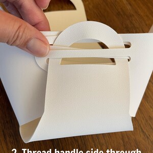May include: A white paper bag with a handle. The handle is being threaded through a slot in the bag. The text "2. Thread handle side through" is visible at the bottom of the image.
