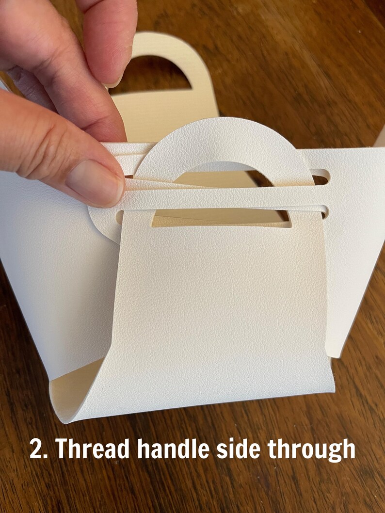 May include: A white paper bag with a handle. The handle is being threaded through a slot in the bag. The text "2. Thread handle side through" is visible at the bottom of the image.