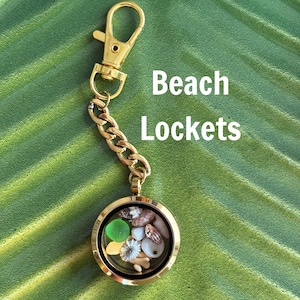 Puede incluir: Un relicario dorado con cadena y cierre, lleno de tesoros con temática de playa. El relicario contiene vidrio de mar, conchas y pequeñas piedras. Las palabras "Beach Lockets" están impresas en blanco.