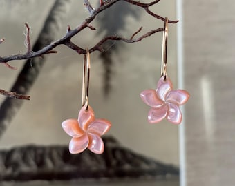 Puede incluir: Un par de pendientes colgantes con dijes de flores de cinco pétalos tallados en material de concha iridiscente de color rosa claro. Los colgantes florales están suspendidos de ganchos de metal dorado, delgados y sencillos.