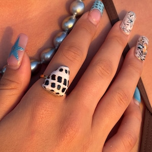 May include: A gold ring with a heart-shaped shell, patterned in black and white. The hand displays long, painted nails with diverse designs, including a blue star, floral patterns, and a tiger face. A pearl necklace is also visible.