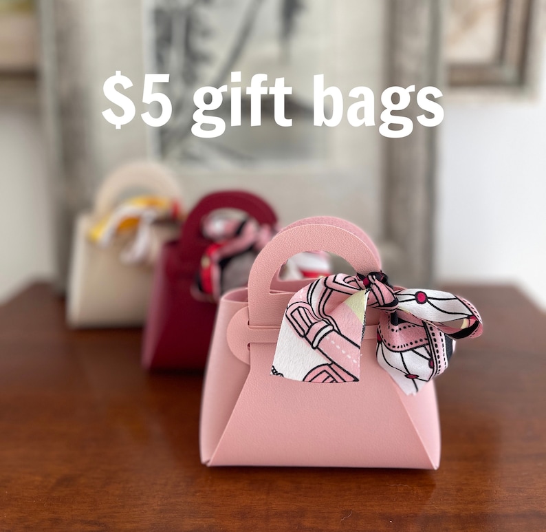 May include: Three gift bags in different colors: beige, burgundy, and pink. Each bag has a ribbon tied around the handle. The pink bag has a patterned ribbon with pink, white, and black colors. The text "$5 gift bags" is visible at the top of the image.