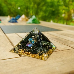 Könnte beinhalten: Eine schwarze und goldene pyramidenförmige Kristall mit eingebetteten Kristallen. Die Pyramide steht auf einer Holzoberfläche.