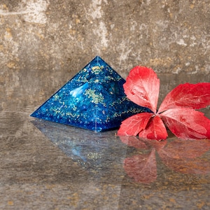 Könnte beinhalten: Ein durchscheinendes blaues Pyramidenobjekt mit eingebetteten goldfarbenen Sprenkeln und kleinen blauen Steinfragmenten, platziert neben einem einzelnen leuchtend roten Blatt mit fünf ausgeprägten Lappen, auf einer reflektierenden grauen Oberfläche.