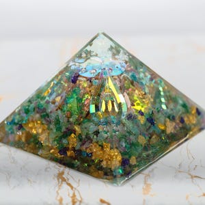 Puede incluir: Una pir&aacute;mide de resina transparente que contiene una mezcla densa de peque&ntilde;os fragmentos de piedra verdes, azules y morados, escamas met&aacute;licas doradas y un elemento central de cristal facetado iridiscente que refleja la luz.