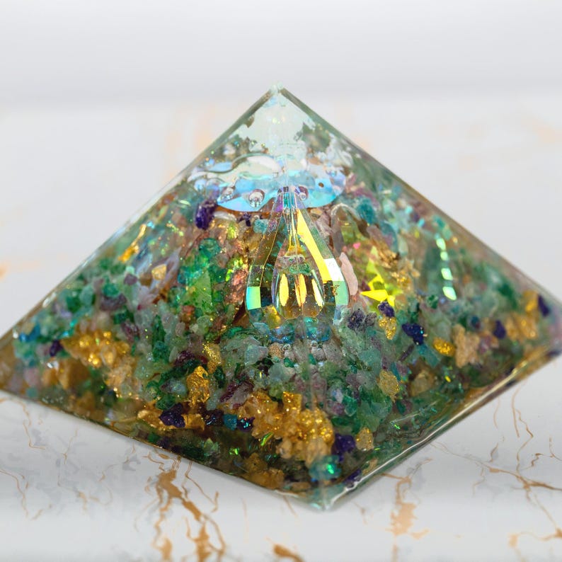 Puede incluir: Una pir&aacute;mide de resina transparente que contiene una mezcla densa de peque&ntilde;os fragmentos de piedra verdes, azules y morados, escamas met&aacute;licas doradas y un elemento central de cristal facetado iridiscente que refleja la luz.