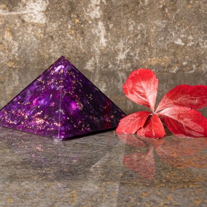 Könnte beinhalten: Eine violette Pyramide mit goldenen Flocken steht auf einer grauen Oberfläche neben einem einzelnen roten Blatt.