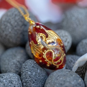 Könnte beinhalten: Ein goldener Drachenanhänger mit roten und goldenen Akzenten ist in einem klaren, roten Glaseier eingeschlossen. Der Anhänger hängt an einer goldenen Kette und liegt auf einem Bett aus grauen Steinen.