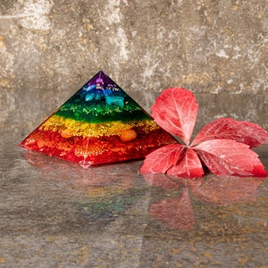 Pode incluir: Uma pirâmide de cristal com cores do arco-íris com uma folha vermelha sobre um fundo cinza. A pirâmide é feita de resina transparente e contém vários cristais, incluindo ametista, citrino e quartzo transparente.
