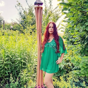Op de afbeelding: Een vrouw in een groene jurk staat in een tuin naast een hoog, koperkleurig beeld met een paarse glazen bol aan de basis. Het beeld heeft een piramidevormige bovenkant.