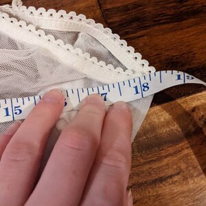Puede incluir: Tela de encaje blanca con una cinta m&eacute;trica blanca que muestra 18 pulgadas. La tela tiene un borde festoneado.