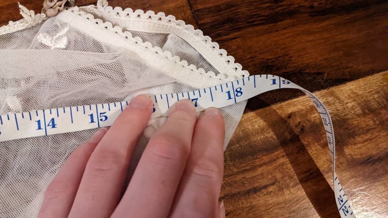 Puede incluir: Tela de encaje blanca con una cinta m&eacute;trica blanca que muestra 18 pulgadas. La tela tiene un borde festoneado.