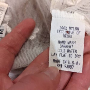Puede incluir: Top de encaje blanco con una etiqueta blanca que dice "100% Nylon Exclusive of Trims, Hand Wash Garment Cold Water Lay Flat to Dry Made in U.S.A. RN# 93087". La etiqueta tambi&eacute;n tiene el nombre de la marca "Claire Pettibon" y la talla "M" escrita en ella.