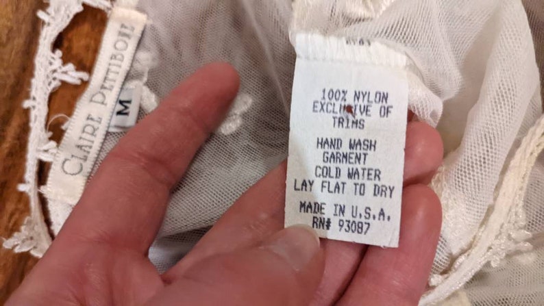Puede incluir: Top de encaje blanco con una etiqueta blanca que dice "100% Nylon Exclusive of Trims, Hand Wash Garment Cold Water Lay Flat to Dry Made in U.S.A. RN# 93087". La etiqueta tambi&eacute;n tiene el nombre de la marca "Claire Pettibon" y la talla "M" escrita en ella.