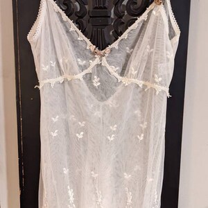 Puede incluir: Un vestido blanco de encaje transparente con delicado bordado floral y ribete de encaje. El vestido tiene un escote redondo y tirantes finos.